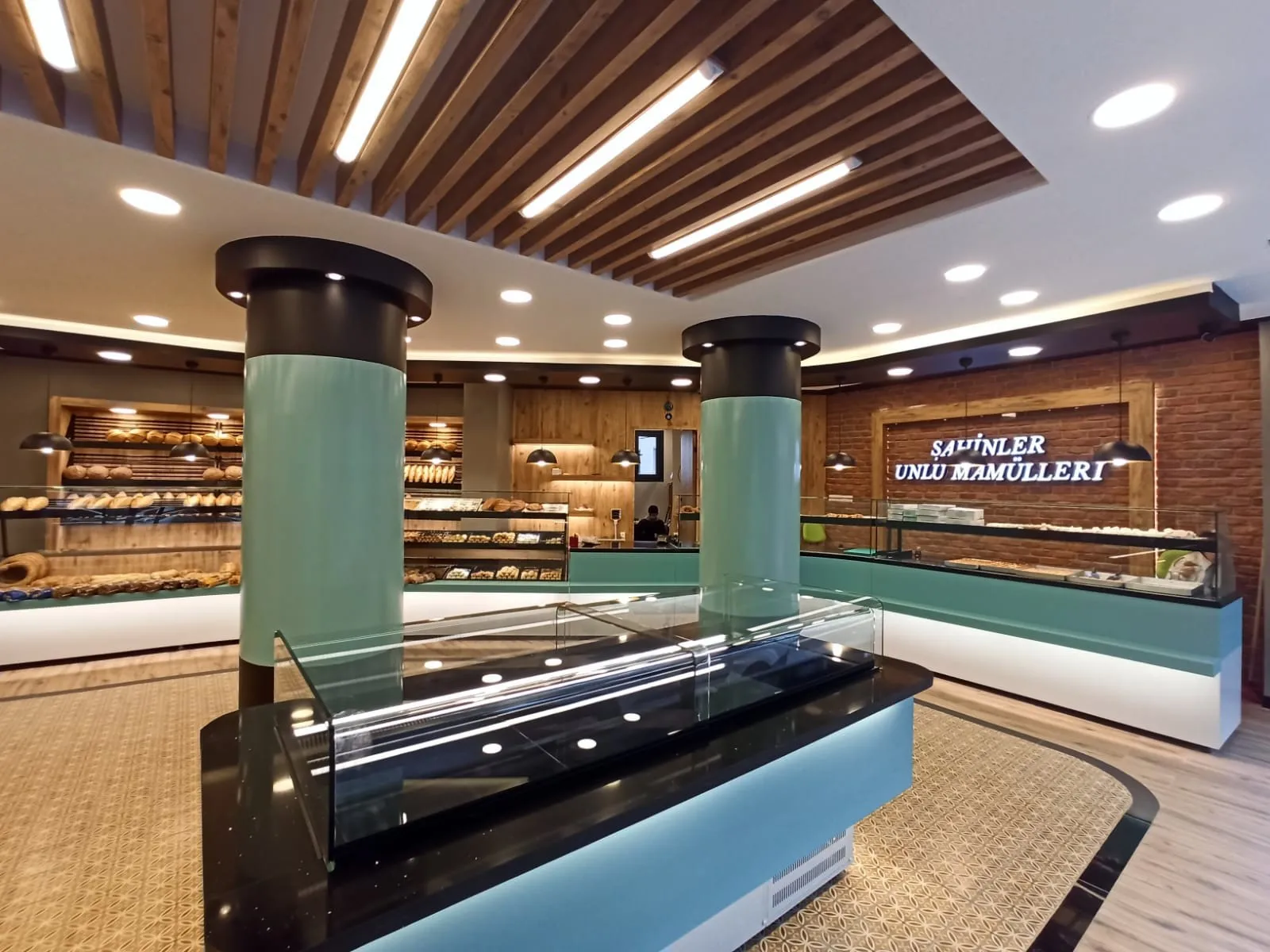 Turkuaz kaplı kolonlar, cam teşhir adası ve ahşap fonda Şahinler tabelasıyla satış alanı