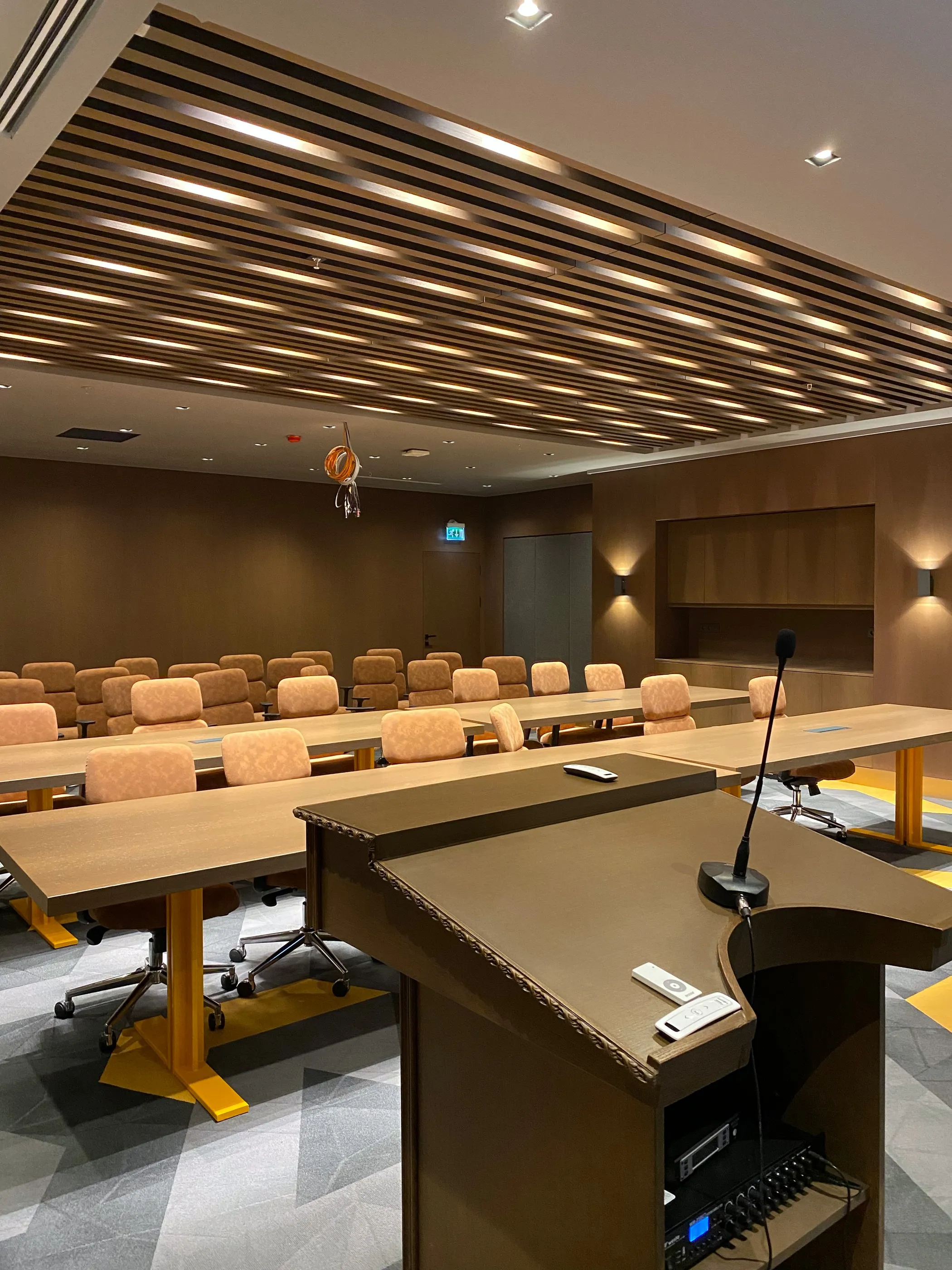 Konferans salonunda ahşap konuşma kürsüsü, uzun toplantı masaları ve sarı-gri desenli halı