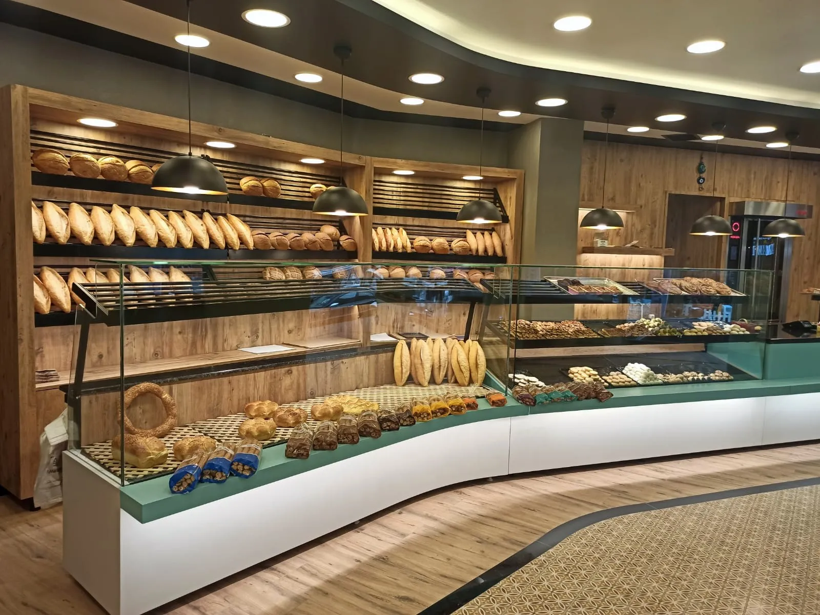 Sarkıt lambaların aydınlattığı ahşap ekmek rafları ve kavisli cam vitrin